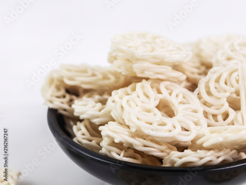 Close-up side view of a black bowl piled high with raw white kerupuk udang (Indonesian prawn crackers) against a clean white background, showcasing their detailed textures and spiral shapes.