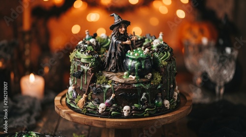 Intricately decorated Halloween chocolate cake with a witch figurine, bubbling green cauldron, skulls, and moss accents on a rustic wooden stand.