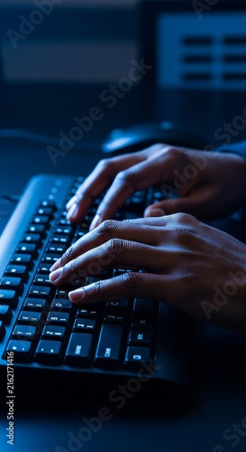 Close up of dark-skinned hands resting on a keyboard, illuminated by a harsh monitor glow in a dark, monotonous and cold corporate office, overtime, mood, weary