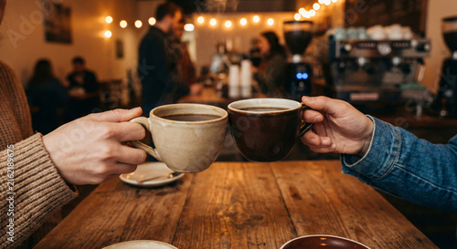 Two people cheering with coffee mugs in a cozy coffee shop during daytime with warm interior lighting