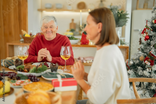 Принти на полотні Senior man smiling during Christmas family dinner at home, enjoying conversation, festive food and meaningful holiday time with family beside Christmas tree