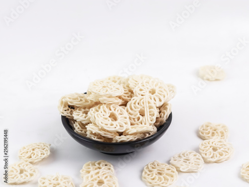 Studio product shot of a black bowl heaped with raw, uncooked white kerupuk udang (traditional Indonesian prawn crackers) alongside loose crackers scattered on a bright white surface.