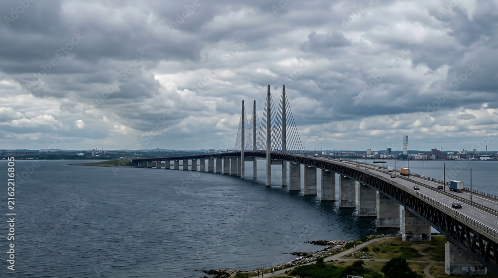Fototapeta premium Long suspension bridge spanning wide water body under cloudy sky.