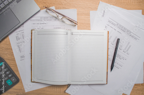 Top view of an open blank notebook surrounded by financial reports, calculator, eyeglasses, laptop and pen on a wooden desk. Business planning and accounting concept. Copy space