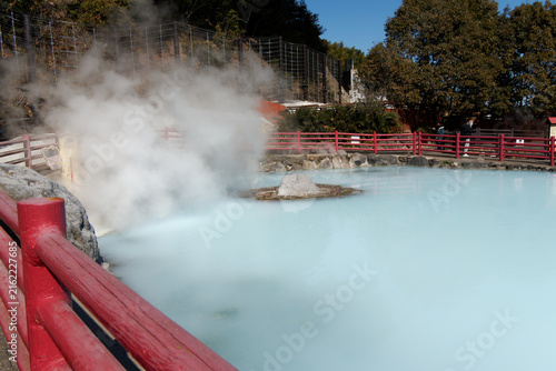 фотографія Wide angle view of a vibrant milky blue geothermal hot spring pool with steam rising and red wooden railing in Beppu