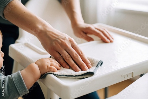 Mother cleans baby highchair tray while child grabs cloth. Parent wipes food mess, toddler interferes playfully. Daily home routine, bonding moment.