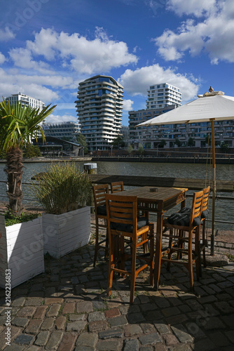 Luxury outdoor restaurant terrace by the river with modern city skyline. Contemporary waterfront cafe patio with wooden furniture and umbrella under blue cloudy sky.