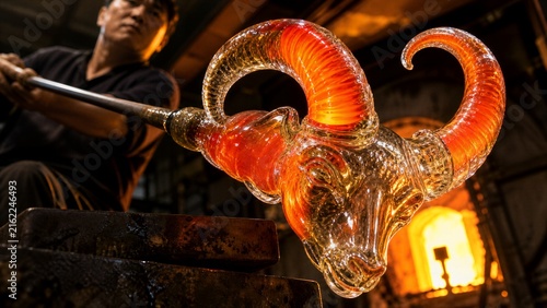 Master Artisan: A craftsman expertly shapes molten glass into a striking ram's head sculpture, the fiery orange glass glowing intensely under the heat of the furnace.