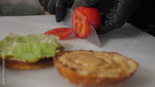 A close-up shot of the chef cutting a tomato for a burger in the restaurant's kitchen, showing the process of preparing a juicy burger. The burger bun is topped with a sauce and lettuce leaves.