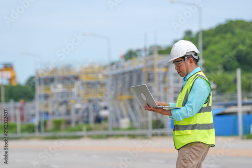 Engineer worker helmet safety vest laptop industrial site inspection, focused male professional using computer near refinery structure during outdoor maintenance work