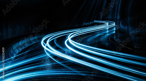 Fotografie Abstract long exposure photograph of luminous electric-blue light ribbons bending gracefully across a slick, shadowed street, evoking relentless forward motion