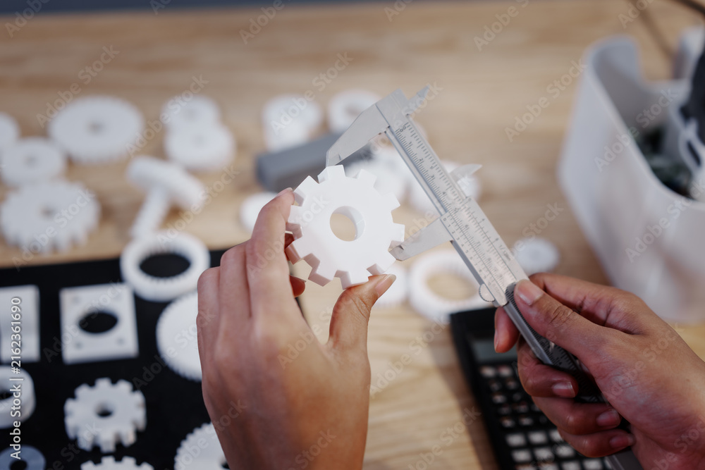 Obraz premium Hands measuring a white plastic gear with calipers on a workbench, with other parts and tools blurred in the background