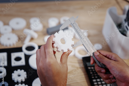 Hands measuring a white plastic gear with calipers on a workbench, with other parts and tools blurred in the background