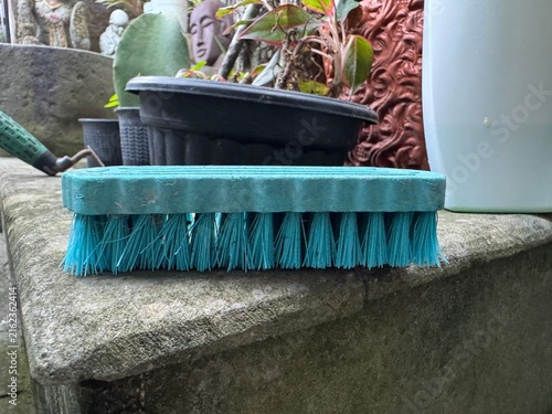 Blue scrub brush with stiff bristles on a weathered stone surface, surrounded by gardening tools and plants.