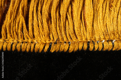 Macro detail of golden yellow yarn fringe stitched along the edge of black textile fabric. Soft fibers, decorative trim and contrast colors emphasize texture.