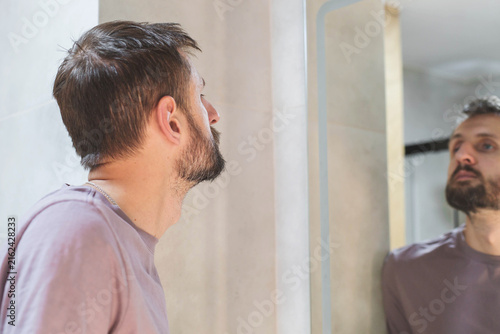 Photography Bearded adult man looking at his reflection in bathroom mirror at home, checking