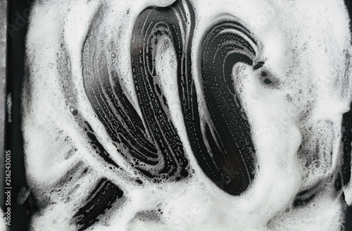 White foam and soap on dark background. The concept of washing oily pan or black car. Abstract pattern.