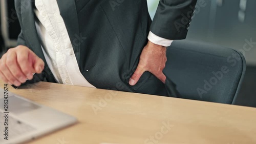 Professional suffers lower back pain while seated at office desk, indicating discomfort from prolonged work. Stress and poor posture contribute to physical strain.