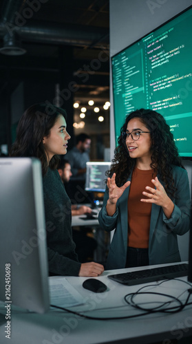 Two women in office discuss code on large screen, symbolizing teamwork, tech, and innovation