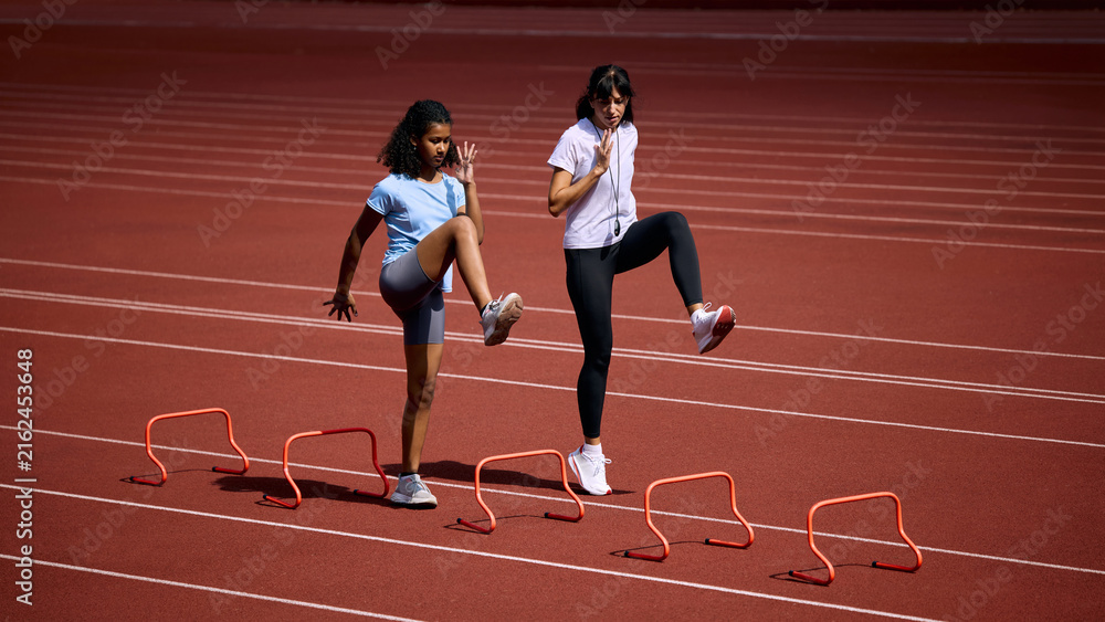 Fototapeta premium Teenage female runner practicing high knee hurdle drill beside adult female coach. Concept of agility and speed training, junior hurdle practice, summer sport coaching.