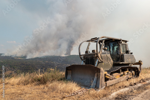 Bulldozer posicionado num terreno acidentado na abertura de faixas de contenção para impedir o avanço de um incêndio florestal massivo que gera uma imensa coluna de fumo no horizonte arborizado