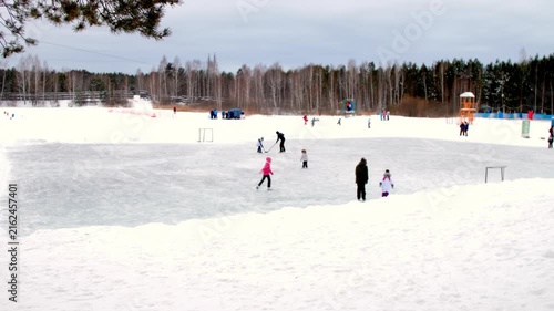 Children skate on ice rink in winter. Kids and parents play hockey and skate on ice rink with solid ice in park against dense forest