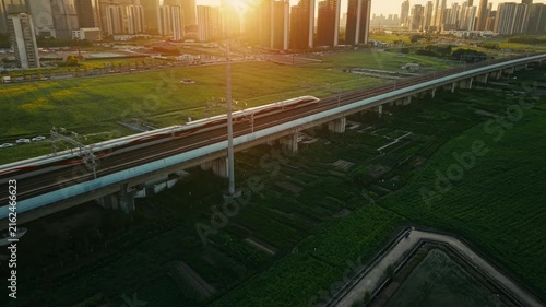 Aerial view of a high speed train travels on an elevated track past tall apartment buildings in a city suburb during sunset in China