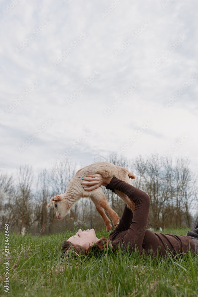 Fototapeta premium Young woman lies on grass in a brown sweater lifting a baby goat above her with a joyful expression in a natural outdoor setting under cloudy sky