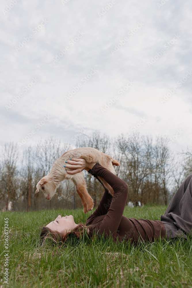 Fototapeta premium Young woman lies on green grass holding a baby goat above her with joy in a natural outdoor setting under cloudy sky