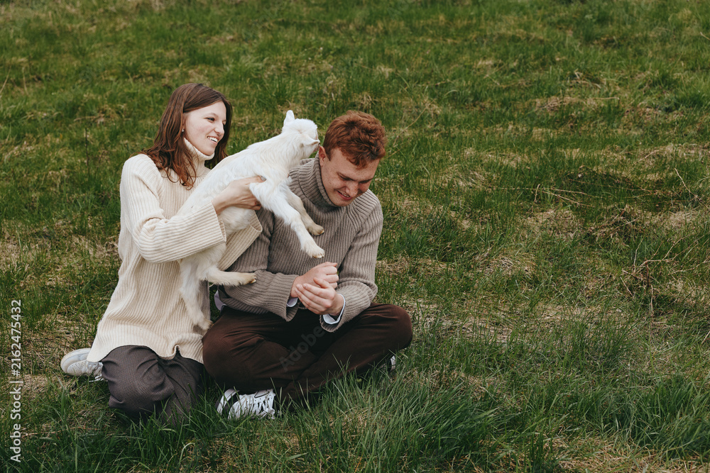 Fototapeta premium Young couple enjoys playful moment with a white baby goat while sitting on green grass in casual sweaters during a relaxed lifestyle outdoor scene.
