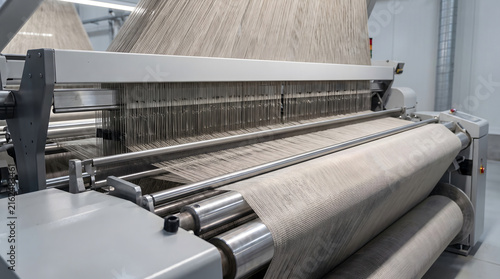 Industrial weaving loom machine processing beige fabric, showing vertical threads feeding into metal rollers and a finished textile roll in a factory