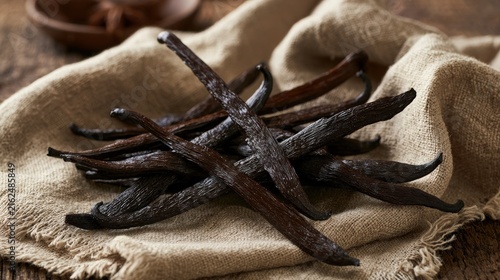 Fragrant, dark brown vanilla pods, a beloved culinary ingredient, are artfully arranged on a textured, light beige linen cloth, with a rustic wooden background hinting at a natural setting.