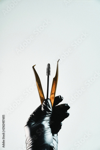 Gold tweezers and mascara wand held together in a gloved han