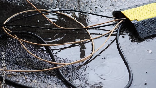 Wet asphalt puddle with tangled black and beige electrical cables leading into yellow black protector. Dangerous outdoor scene combining rain water and live wires creating short circuit hazard 4k