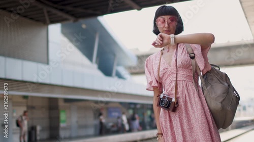 A female traveler with a camera and a backpack stands on a railway station platform. She is checking her watch and waiting for the train. Train travel. A young woman with a camera.