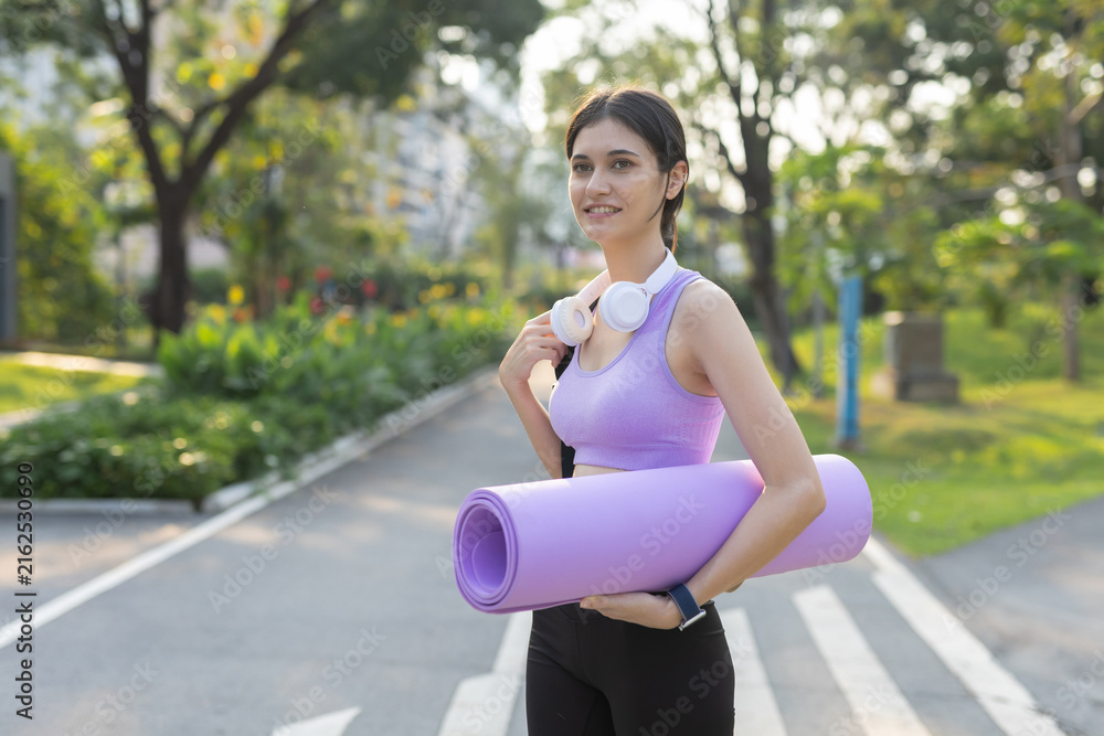 Fototapeta premium Smiling young woman holding yoga mat outdoors in a green park, wearing headphones before fitness training, stretching, yoga practice, exercise, wellness and recreation.