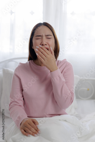 Foto Sleepy woman yawning after waking up in bed, covering her mouth with hand in a bright bedroom, representing morning fatigue, tiredness, drowsiness, sleepiness, bedtime routine and lack of sleep