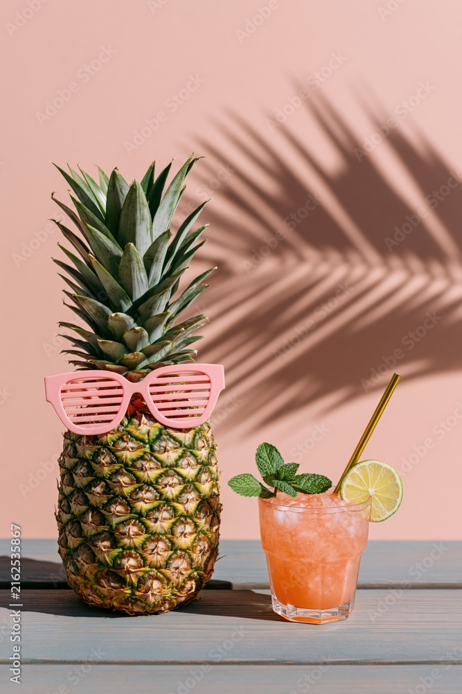 Fototapeta premium Pineapple with pink shutter shades and a lime garnished cocktail casting palm shadow on pastel pink for summer drink poster and backdrop