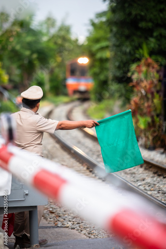 Fotografie Selective focus to Thai railway officer holding a green signal flag at a level crossing safety barrier as a passenger train approaches along the tracks in a lush green environment
