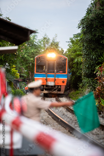 Принти на полотні Selective focus to passenger train with blurry Thai railway officer holding a green signal flag at a level crossing safety barrier as a passenger train approaches along the tracks in a lush green