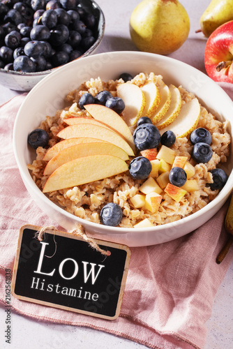 Bowl of oatmeal topped with sliced apple, pear and fresh blueberries, wholesome low histamine and anti inflammatory breakfast with visual aid with text Low Histamine
