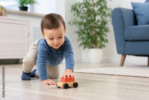 Toddler boy crawling on a wooden floor while playing with a toy car. Happy young child pushing a wooden toy in a living room