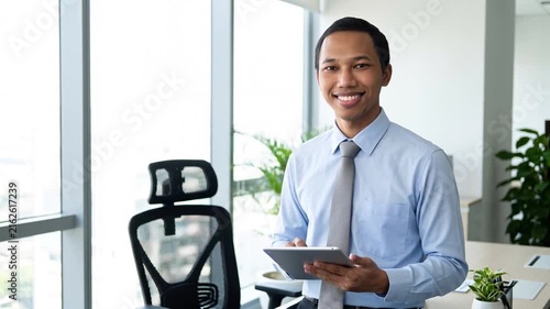 Busy young Indonesian business man executive standing in office using digital tablet. Young professional businessman corporate manager wearing suit and glasses holding tab working on finance project