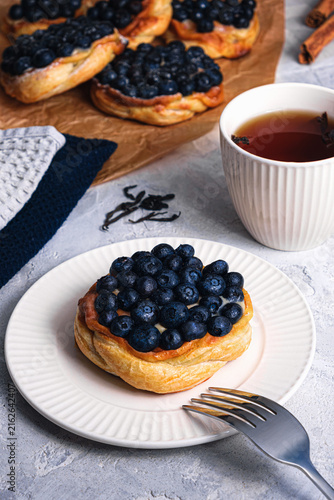 Blueberry puff pastry tart with fork on white plate, tea cup, cinnamon sticks and loose tea leaves on textured table, cozy breakfast scene