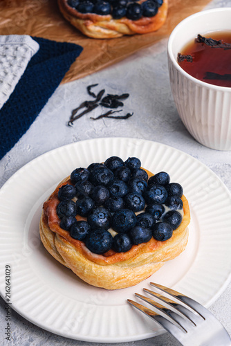 Blueberry puff pastry tart with fork on white plate, tea cup, cinnamon sticks and loose tea leaves on textured table, cozy breakfast scene