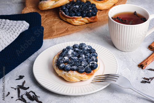 Blueberry puff pastry tart with sugar powder and fork on white plate, tea cup, cinnamon sticks and loose tea leaves on textured table, cozy breakfast scene