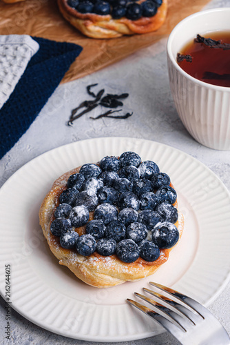 Blueberry puff pastry tart with sugar powder and fork on white plate, tea cup, cinnamon sticks and loose tea leaves on textured table, cozy breakfast scene