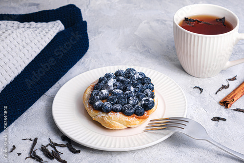 Blueberry puff pastry tart with sugar powder and fork on white plate, tea cup, cinnamon sticks and loose tea leaves on textured table, cozy breakfast scene