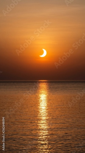 Solar eclipse setting over calm ocean water with golden shimmering path, dramatic sunset sky, crescent sun, seaside landscape, tranquil day