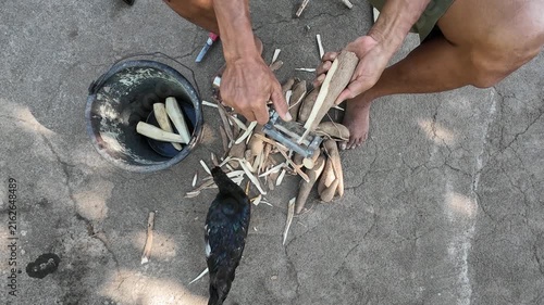 Top view of barefoot farmer peeling fresh cassava roots with traditional handmade peeler on concrete floor preparing tape tapai fermented food with black chickens nearby in Lombok Indonesia